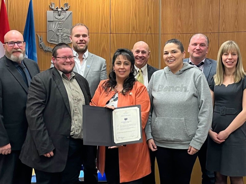 A group of people in a wood paneled room. The woman in the center holds a proclamation.
