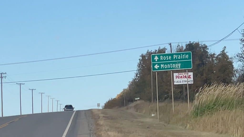 A road with a truck going over the hill in the centre and on the right there is a green direction sign pointing straight for Rose Prairie and left for Montney.