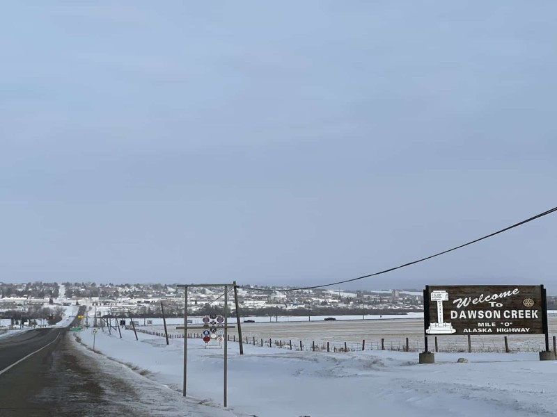 A highway with snow on either side and a wooden sign that reads "Dawson Creek"