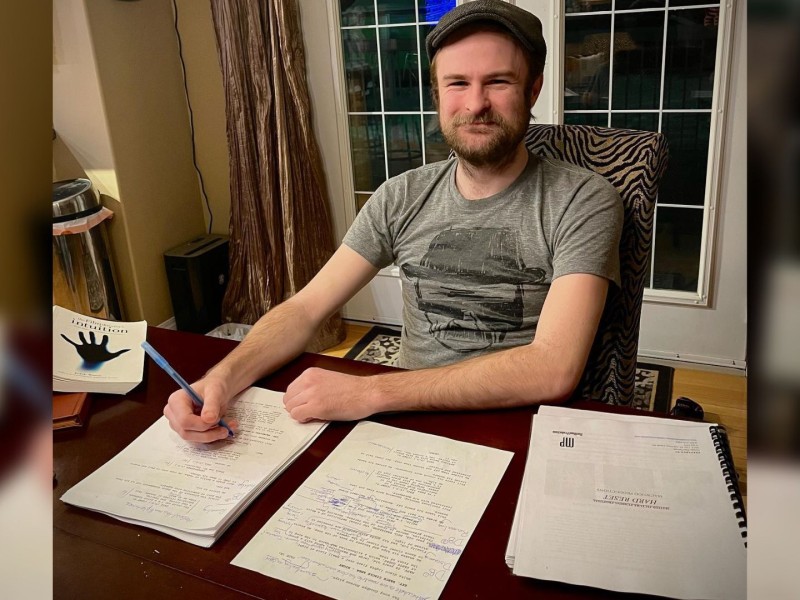 A young man in a grey shirt and cap sits at a table with a script in front of him.