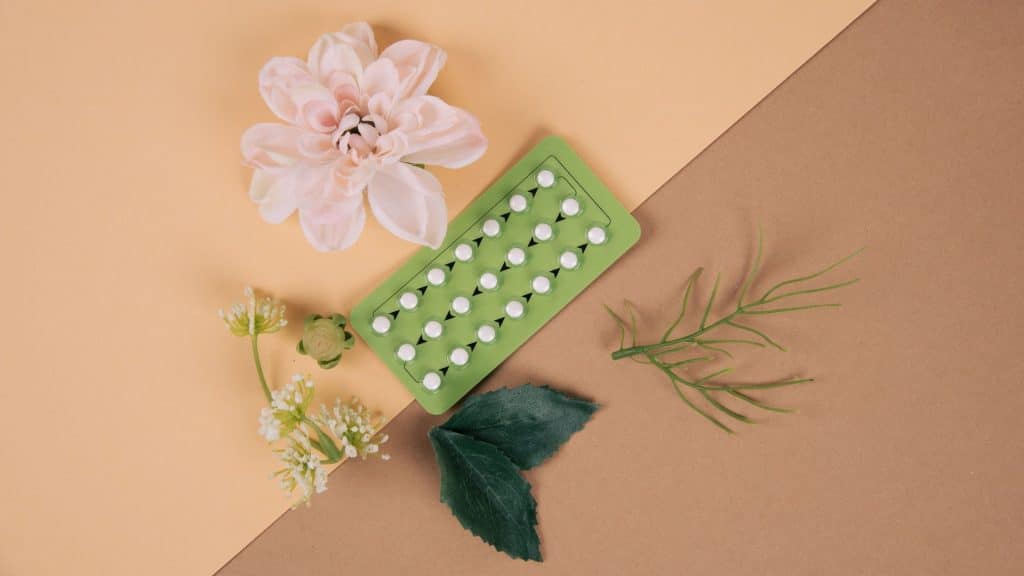 A three week pack of birth control pills surrounded by leaves and flowers on a pink and brown background.