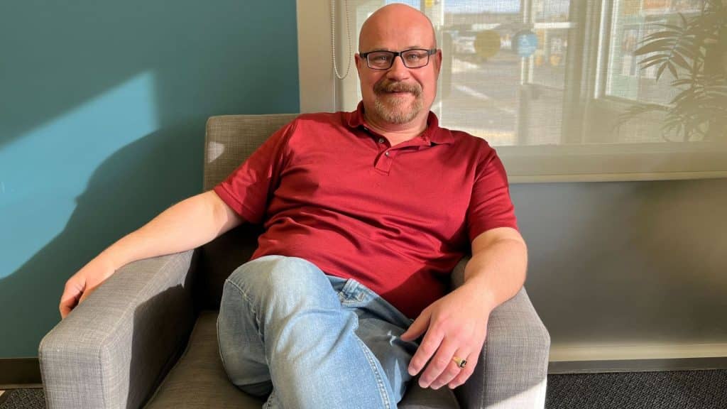 A bald, middle aged man wearing a red shirt and jeans sitting in a grey chair.