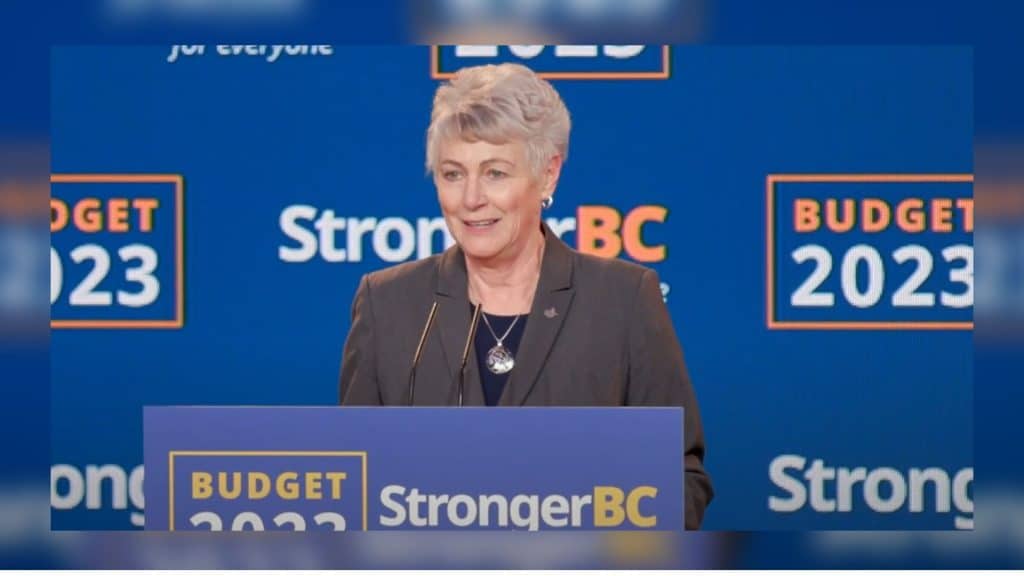 An older woman in a suit stands behind a blue podium in front of a blue background.