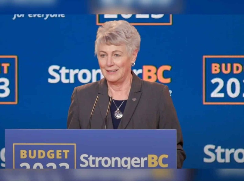An older woman in a suit stands behind a blue podium in front of a blue background.