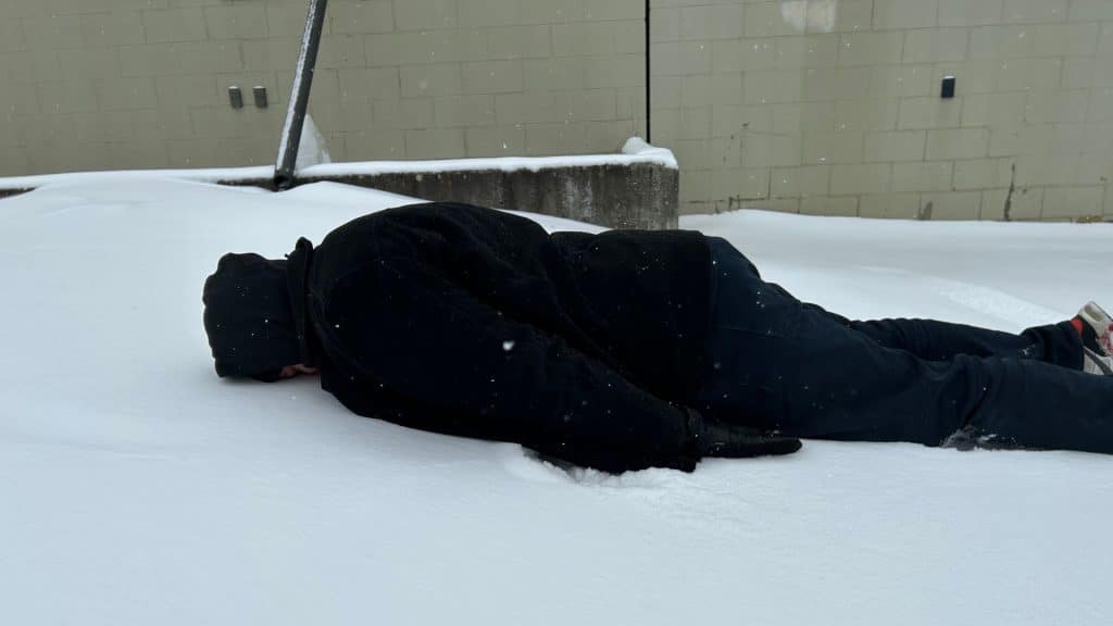 A man in dark clothes laying facedown in the snow in front of a building.