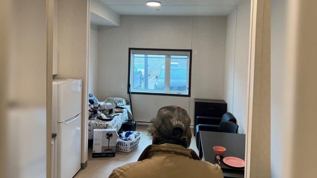 A man with his back to the camera entering a small room with a bed, table, couch and kitchenette.