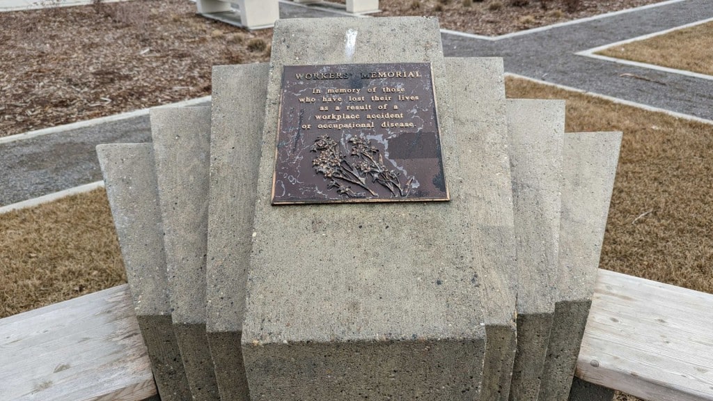 A Workers Memorial in Fort St. John in front of a parking lot full of cars. The plaque reads "Workers Memorial. In memory of those who have lost their lives as a result of a workplace accident or occupational disease."