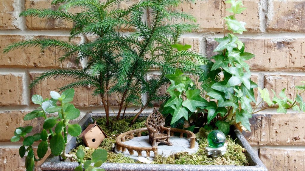 A little house and chair in a small flower pot against a brick wall.