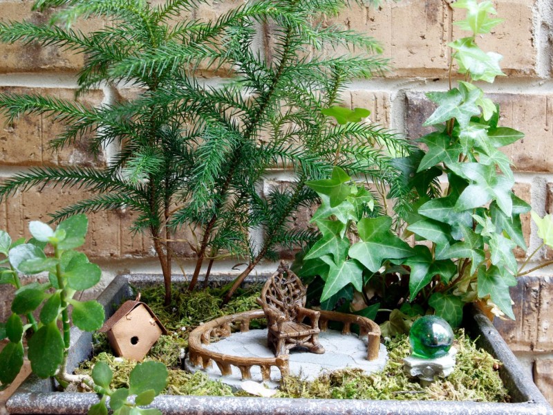 A little house and chair in a small flower pot against a brick wall.