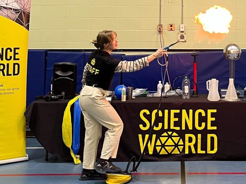 A lady in a black shirt for Science World making a fireball scientifically in front of a Science World table and poster.
