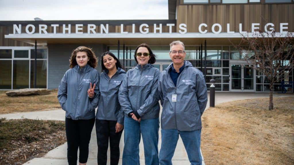 Four people standing in front of Northern Lights College wearing matching grey jackets.