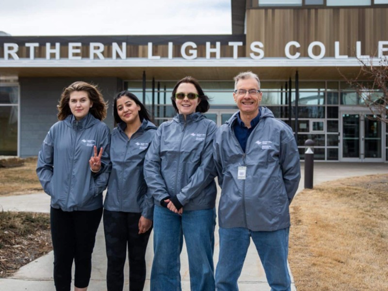 Four people standing in front of Northern Lights College wearing matching grey jackets.