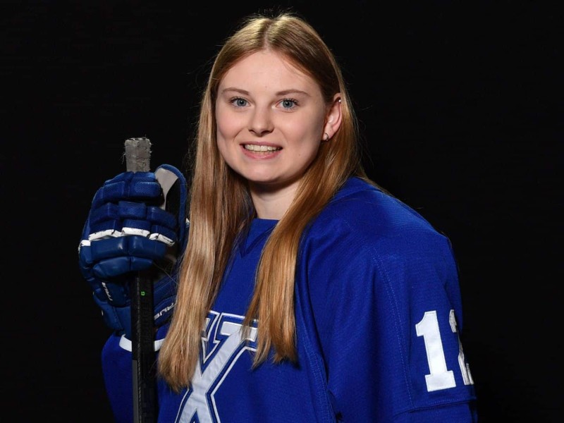 A photo of a blonde haired girl in a hockey jersey holding a hockey stick in front of a black background.