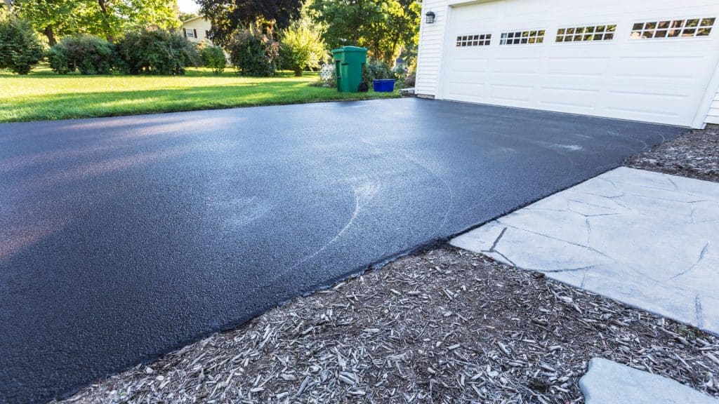 A picture of a driveway in front of a white garage with a lawn beside it and trees in the background.