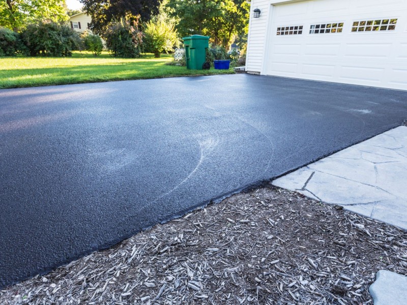 A picture of a driveway in front of a white garage with a lawn beside it and trees in the background.