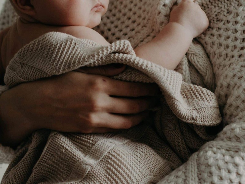 A person in a white knit sweater holding a baby wrapped in a grey knit blanket.