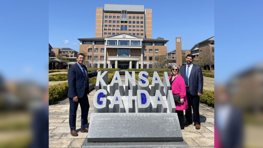 Three people standing beside a large concrete sign that reads "Kansai Gaidai" in front of a large building.