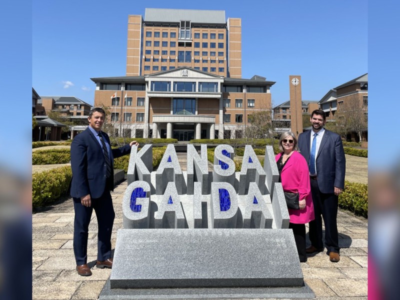 Three people standing beside a large concrete sign that reads "Kansai Gaidai" in front of a large building.