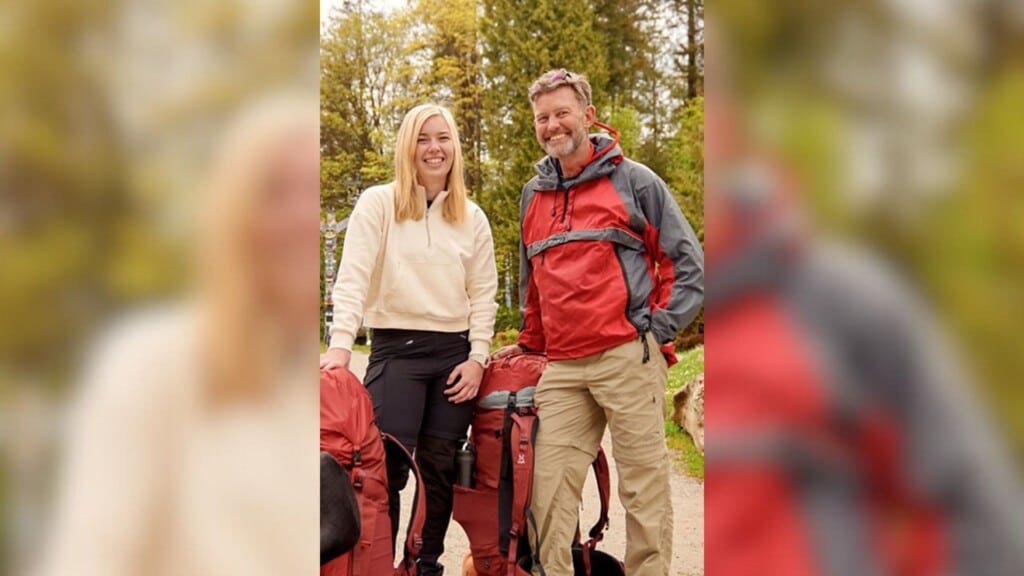 A man in a red jacket standing next to a blonde women in a white sweater in front of some evergreen trees.