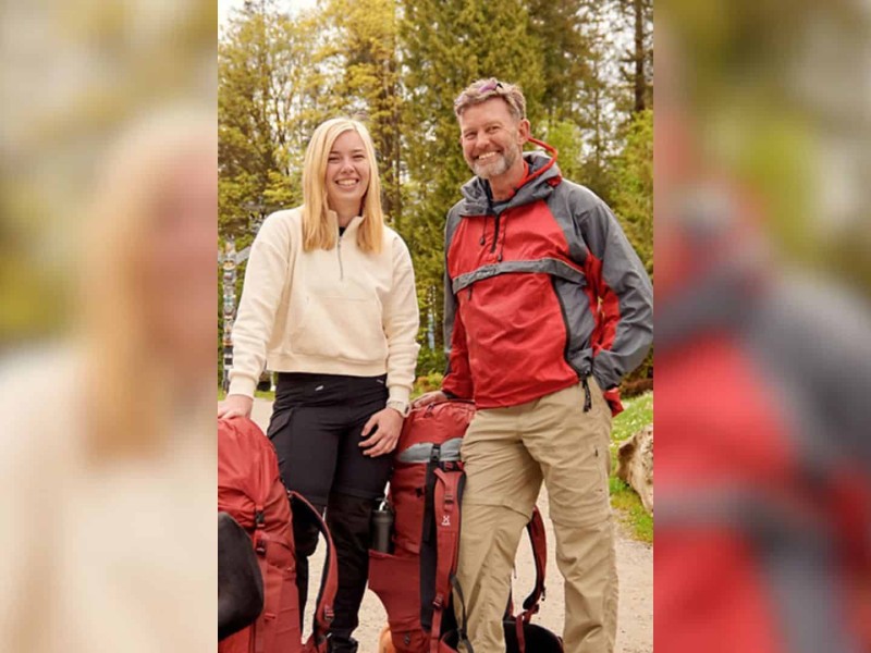 A man in a red jacket standing next to a blonde women in a white sweater in front of some evergreen trees.