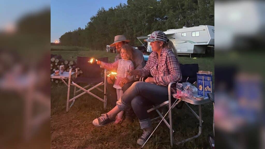 A man and woman sitting in camping chairs while their daughter holds a sparkler in front of her. There is a camper in the background.