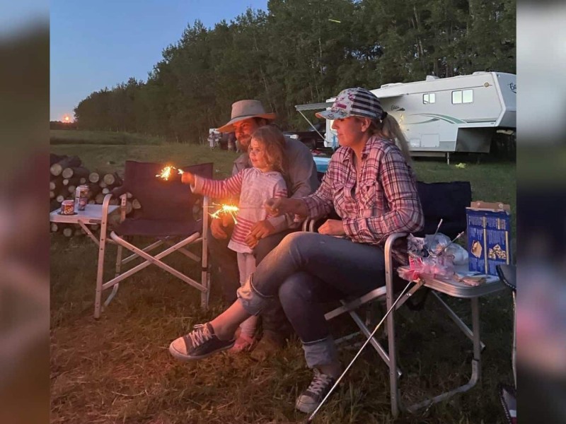 A man and woman sitting in camping chairs while their daughter holds a sparkler in front of her. There is a camper in the background.