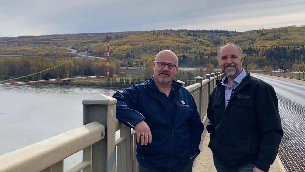Two middle aged men stand on a bridge over a river.