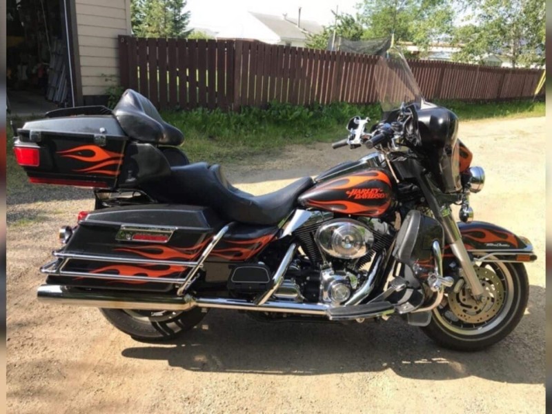 A picture of a black Harley Davidson motorcycle with flames on it on a dirt alley way in front of a brown fence.