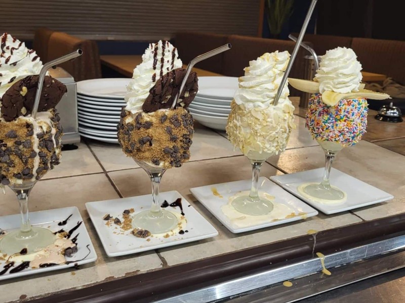 Four very full and over the top milkshakes sitting on plates on a counter in a cafe.