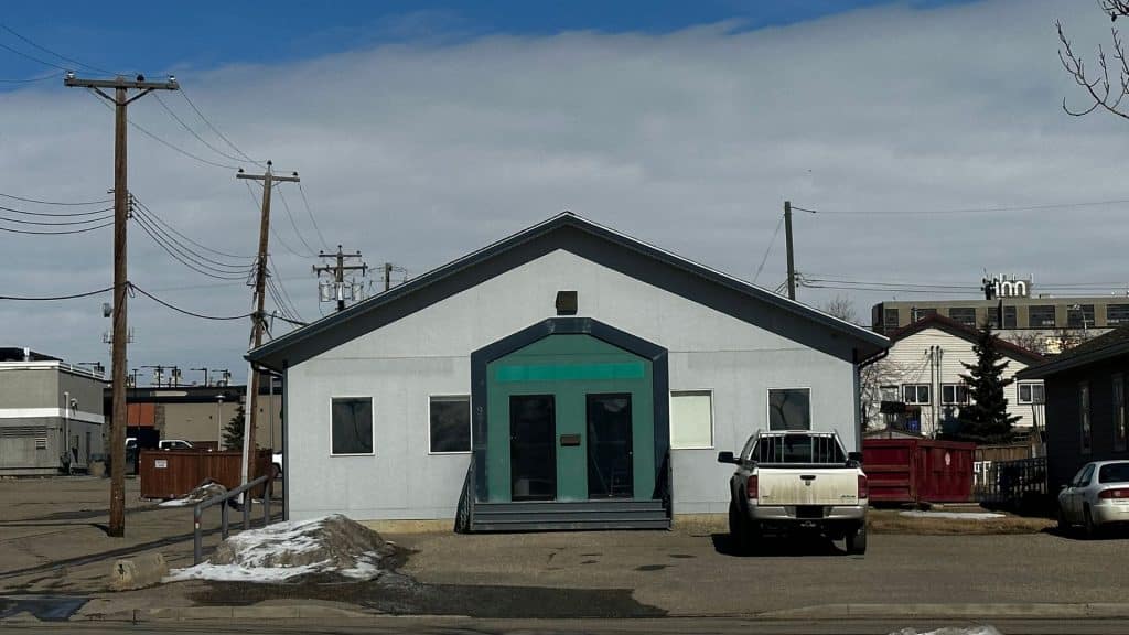 A photo of a grey building with a large green entryway with snow in the parking lot and a white truck out front.
