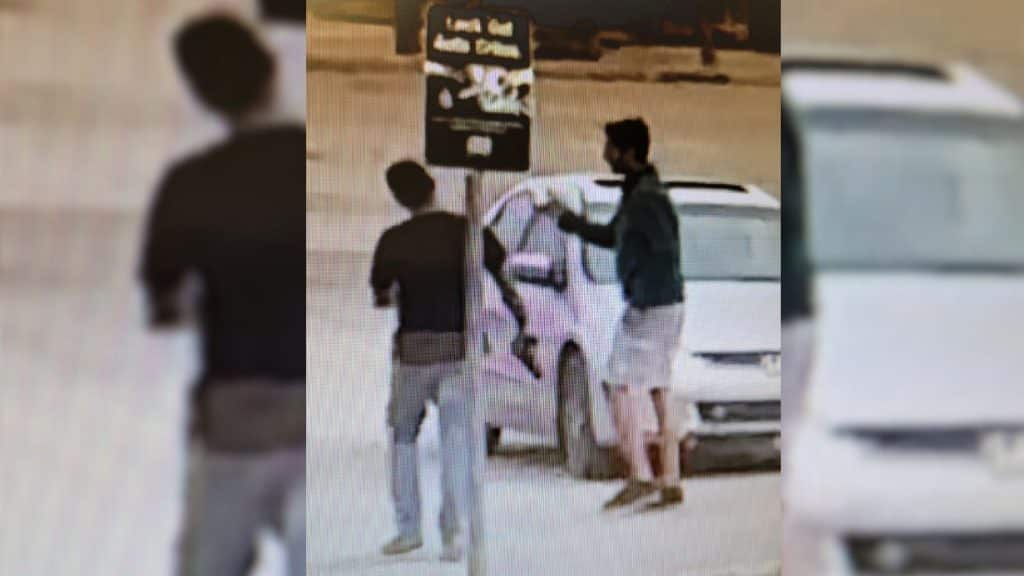 A grainy photo of two men in front of a white car. One of the men is holding what looks like a handgun.