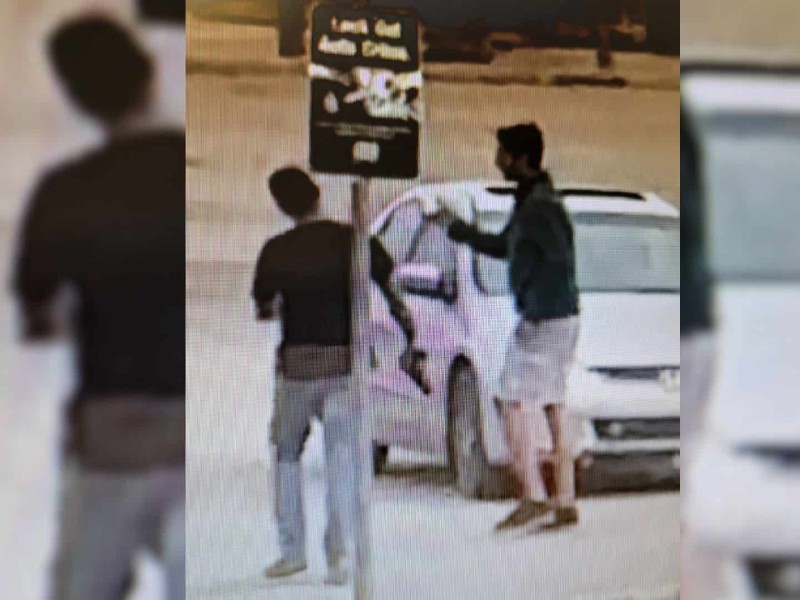 A grainy photo of two men in front of a white car. One of the men is holding what looks like a handgun.