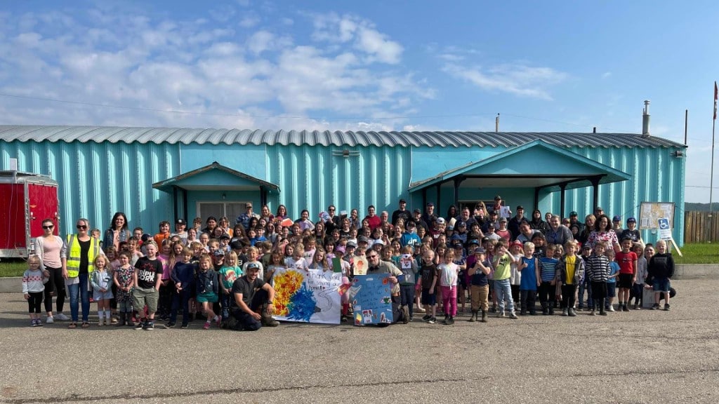 A large group of students, staff and firefighters standing in front of a blue building on a nice day holding a painted sign.