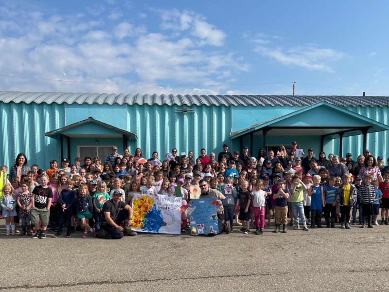 A large group of students, staff and firefighters standing in front of a blue building on a nice day holding a painted sign.