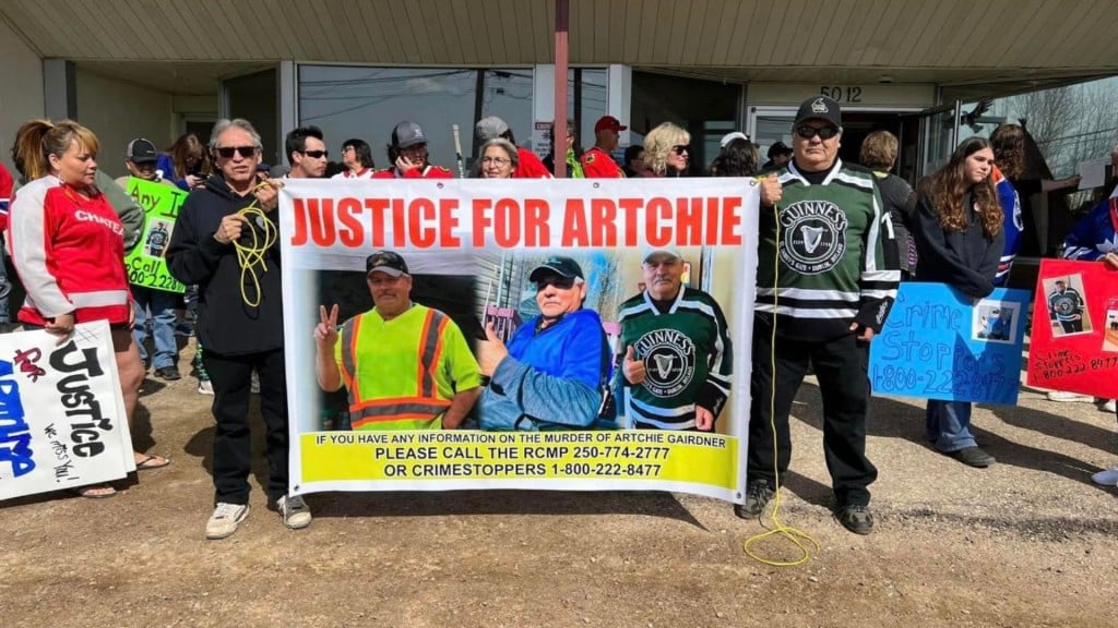 A group of people standing in front of a building, with two men, one in a jersey, holding a sign that says "Justice for Artchie" across the top.