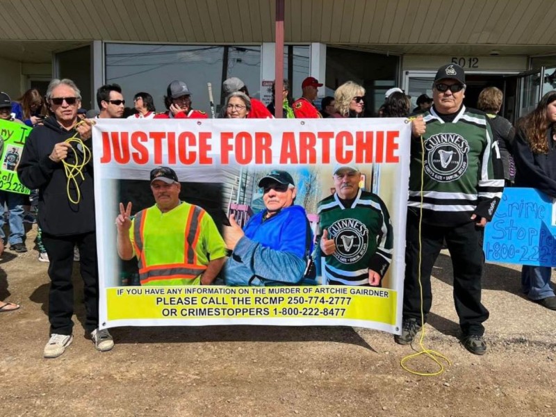 A group of people standing in front of a building, with two men, one in a jersey, holding a sign that says "Justice for Artchie" across the top.