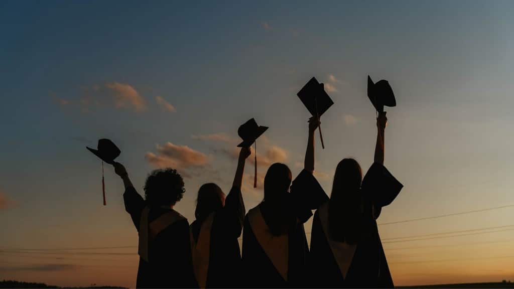 A silhouette of four students holding graduation caps against a sunset.