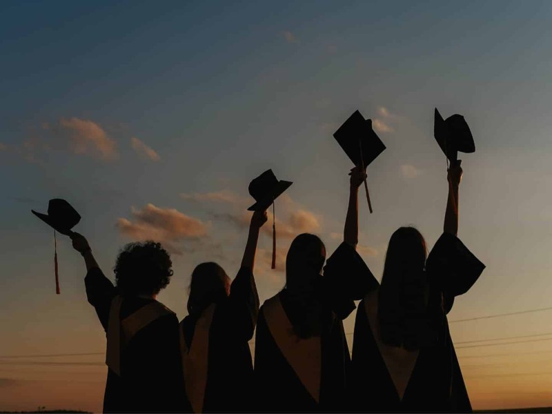 A silhouette of four students holding graduation caps against a sunset.