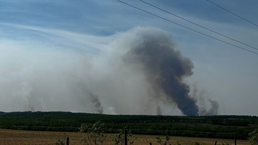 A picture of wildfire smoke behind a hill, a field, a barbed wire fence and some foliage.