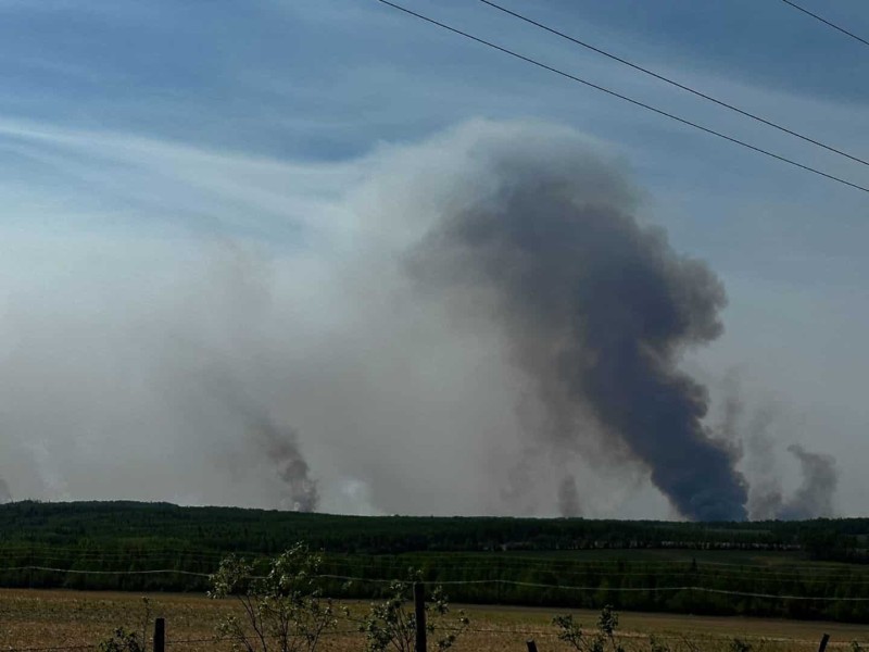 A picture of wildfire smoke behind a hill, a field, a barbed wire fence and some foliage.