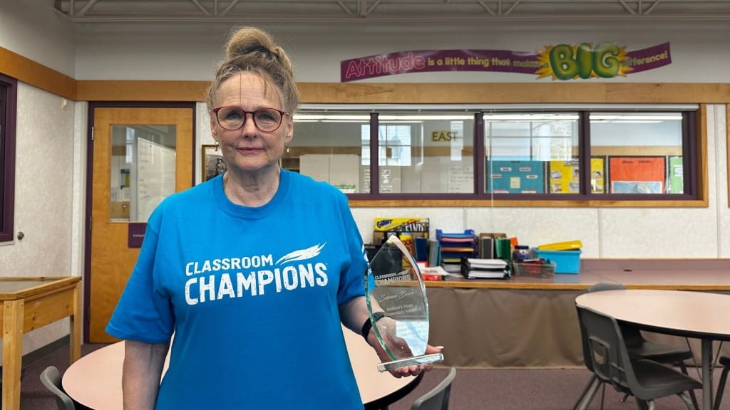 A teacher with her hair pinned up and wearing a blue "Classroom Champions" t-shirt standing in a classroom holding an award.