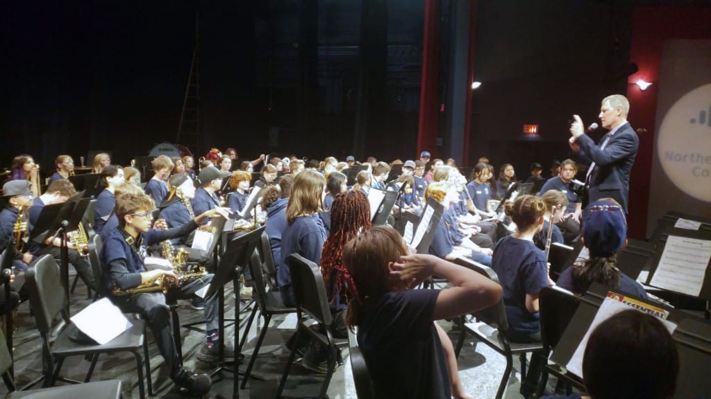 A group shot of band students being talked to by a man at the front of the group with a microphone.