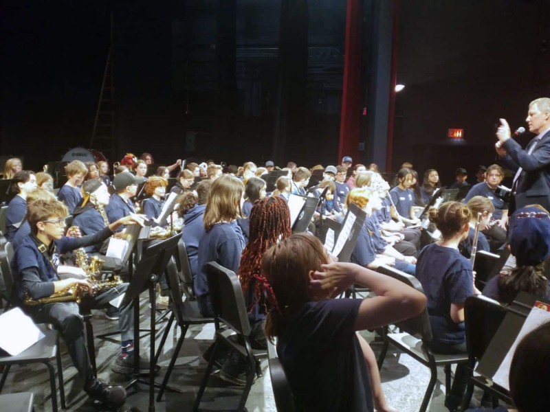 A group shot of band students being talked to by a man at the front of the group with a microphone.