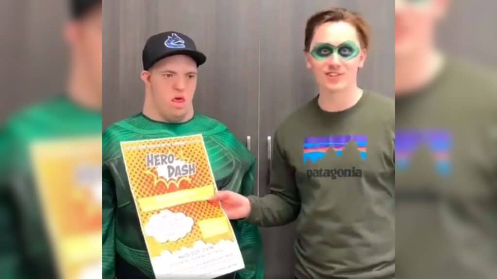 Two men standing next to eachother in front of a brown cupboard, one is wearing a green superhero costume, the other has a green mask painted on. One is holding a "Hero Dash" poster.