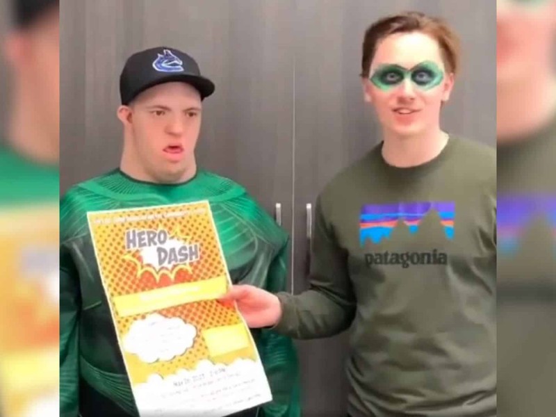 Two men standing next to eachother in front of a brown cupboard, one is wearing a green superhero costume, the other has a green mask painted on. One is holding a "Hero Dash" poster.
