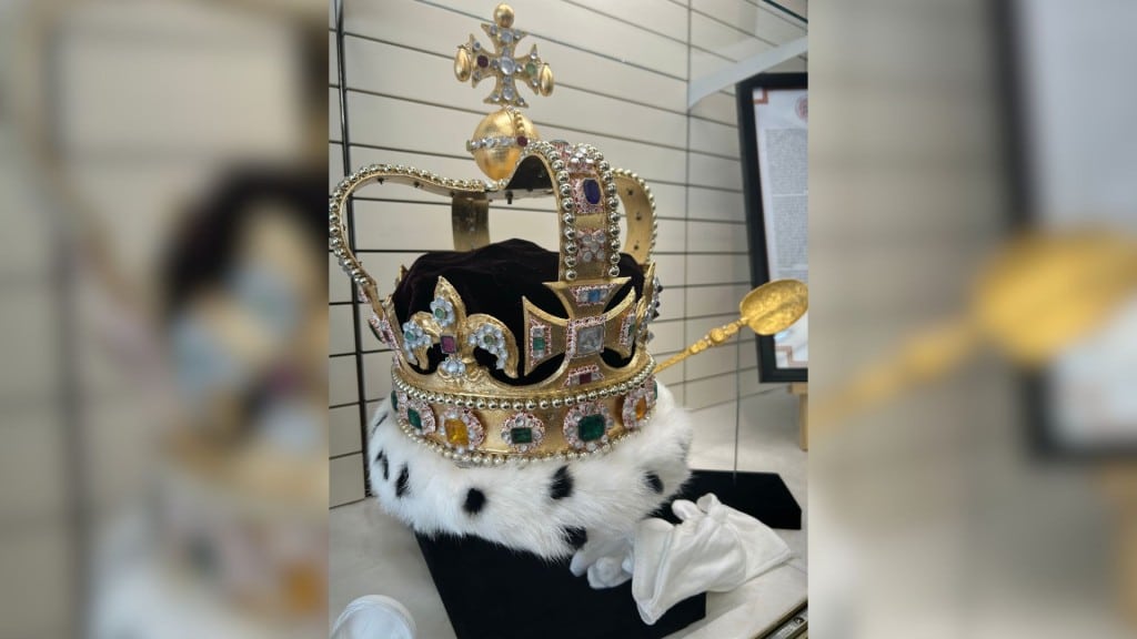 A picture of a replica of the coronation crown and spoon displayed in front of a white wall behind a glass case.