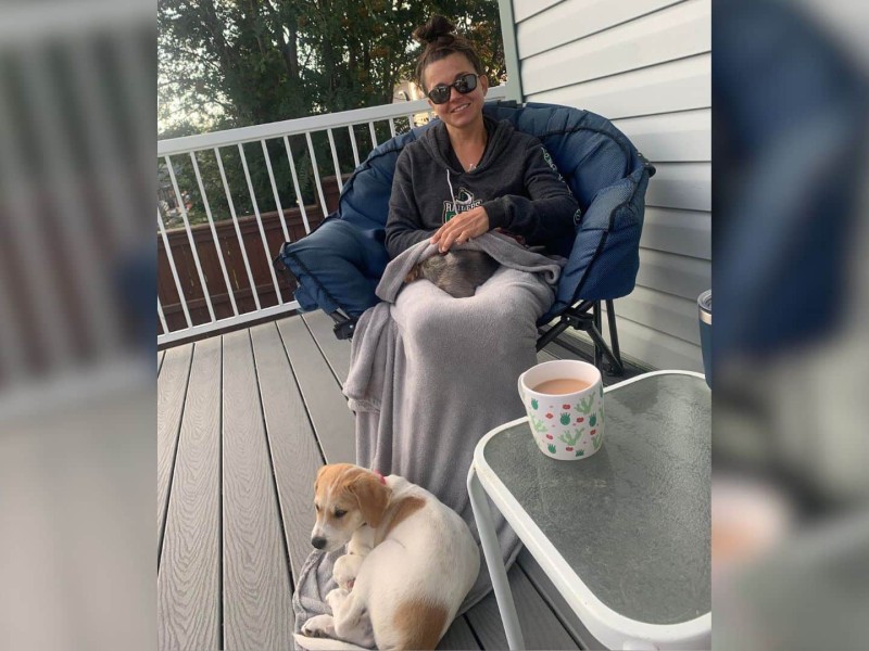 A woman with her hair pinned up and sunglasses on sitting in a camping chair with a blanket on her lap and a dog sitting in front of her.