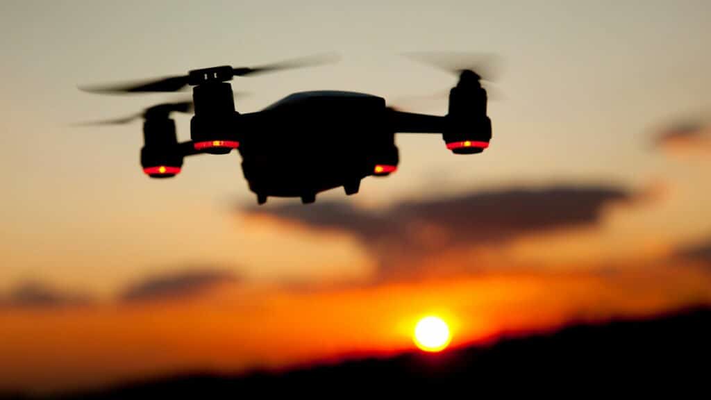 A picture of a black drone with red lights in front of an orange sunset.