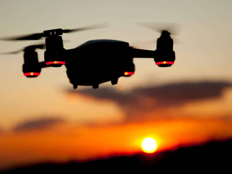 A picture of a black drone with red lights in front of an orange sunset.