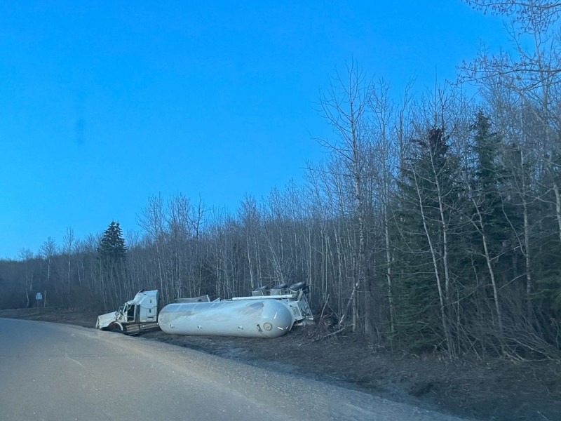 A white tanker truck rolled over on the side of the road next to a forest.